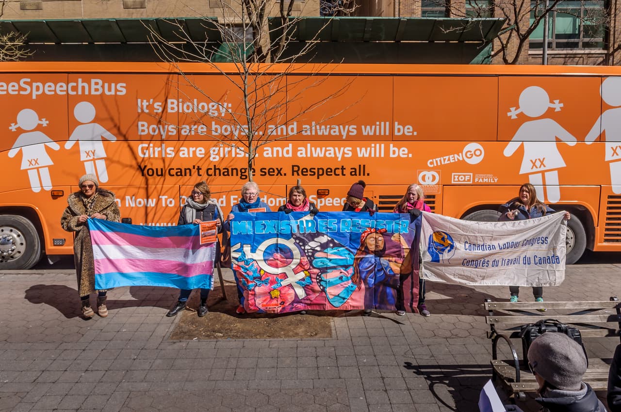 A bus with #FreeSpeechBus stamped onto the side. Anti-LGBTQ protesters stand in front of it with banners