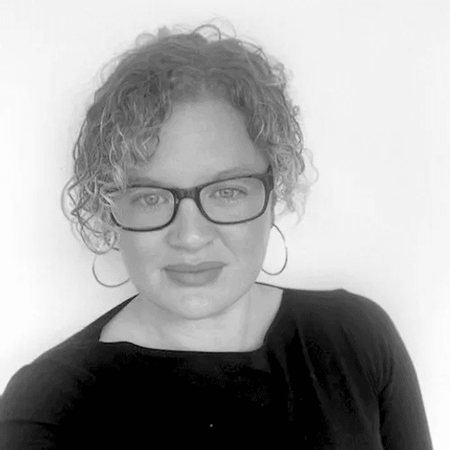 A head and shoulders photograph of a white woman with curly hair tied back, glasses, hoop earrings and dark top