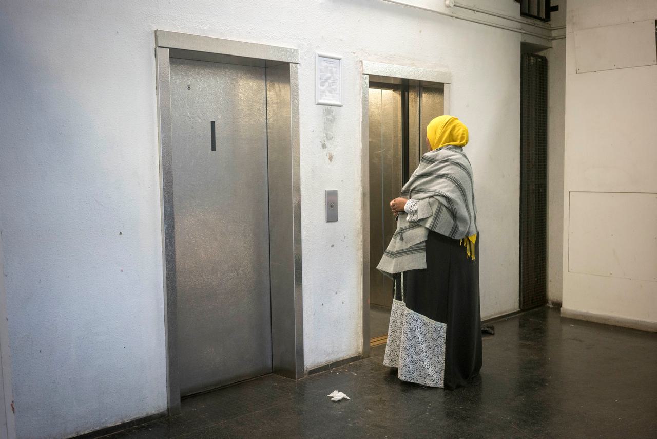 A woman in a yellow headscarf and black and white shawl stands by a bank of lifts on a council estate