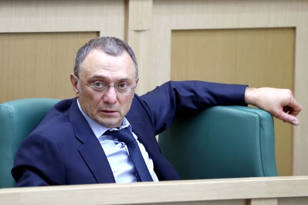 Suleyman Kerimov, a white middle aged man wearing a navy blue suit, sits in a green leather chair in a parliamentary council chamber