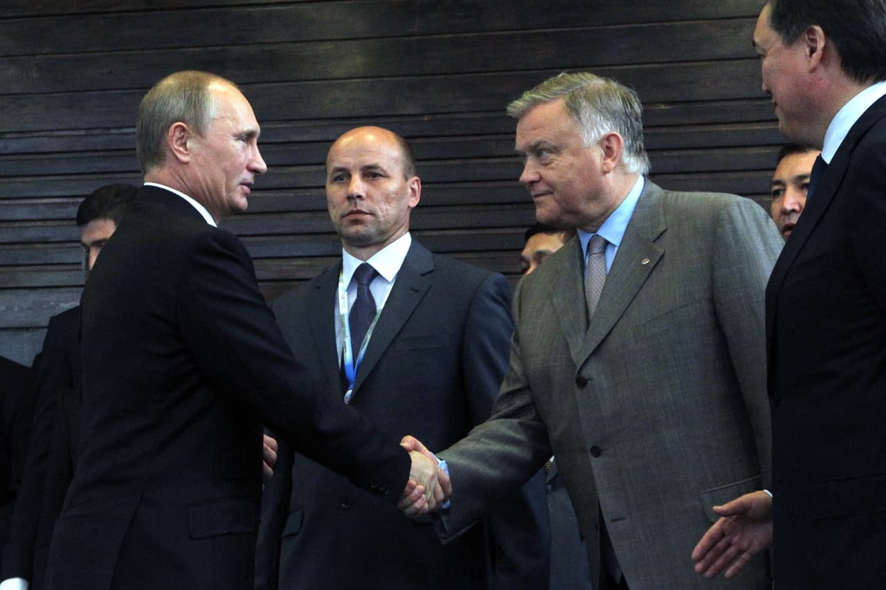 Vladimir Putin, wearing a black suit, shakes hands with a broad-shouldered white man with grey hair. They are surrounded by other men in dark suits