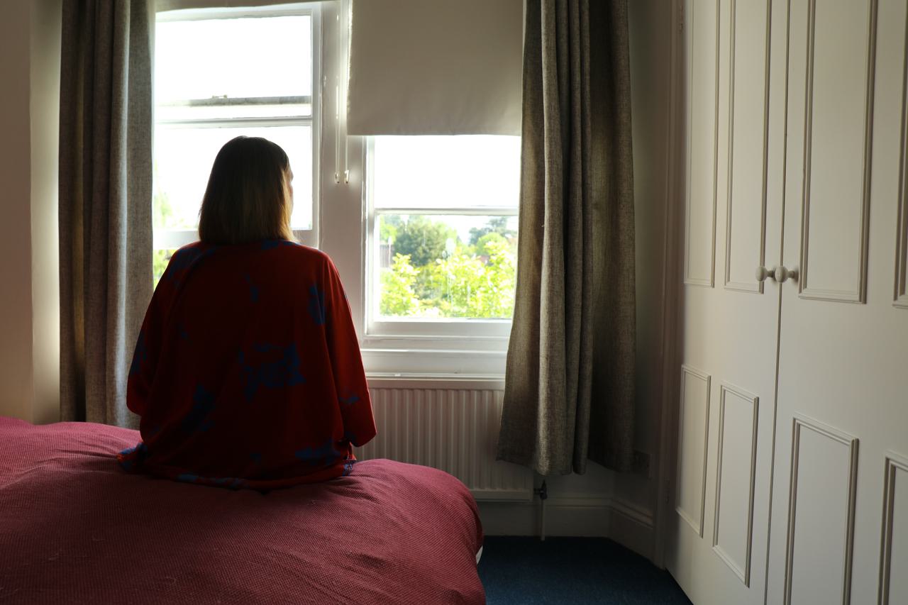 A woman sits on a bed looking out of a window. Her back is to the camera. Trees and sunlight can be seen through the window.