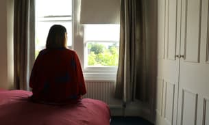 A woman sits on a bed looking out of a window. Her back is to the camera. Trees and sunlight can be seen through the window.