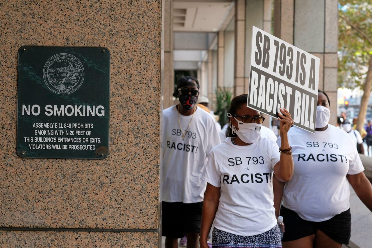 Protestors in white T-shirts with the slogan ‘Say No to SB 793’ walk past a No Smoking sign. One of the protestors carries a sign that says ‘SB 793 is a racist law’