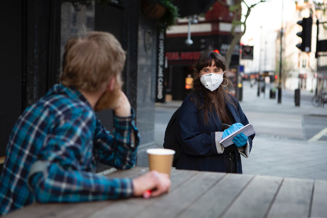 A man in a checked shirt talks to a woman in a protective mask and gloves as she takes notes